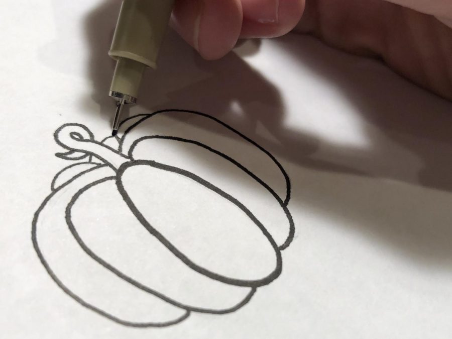 An+artist+uses+a+pen+to+ink+a+small+drawing+of+a+pumpkin.